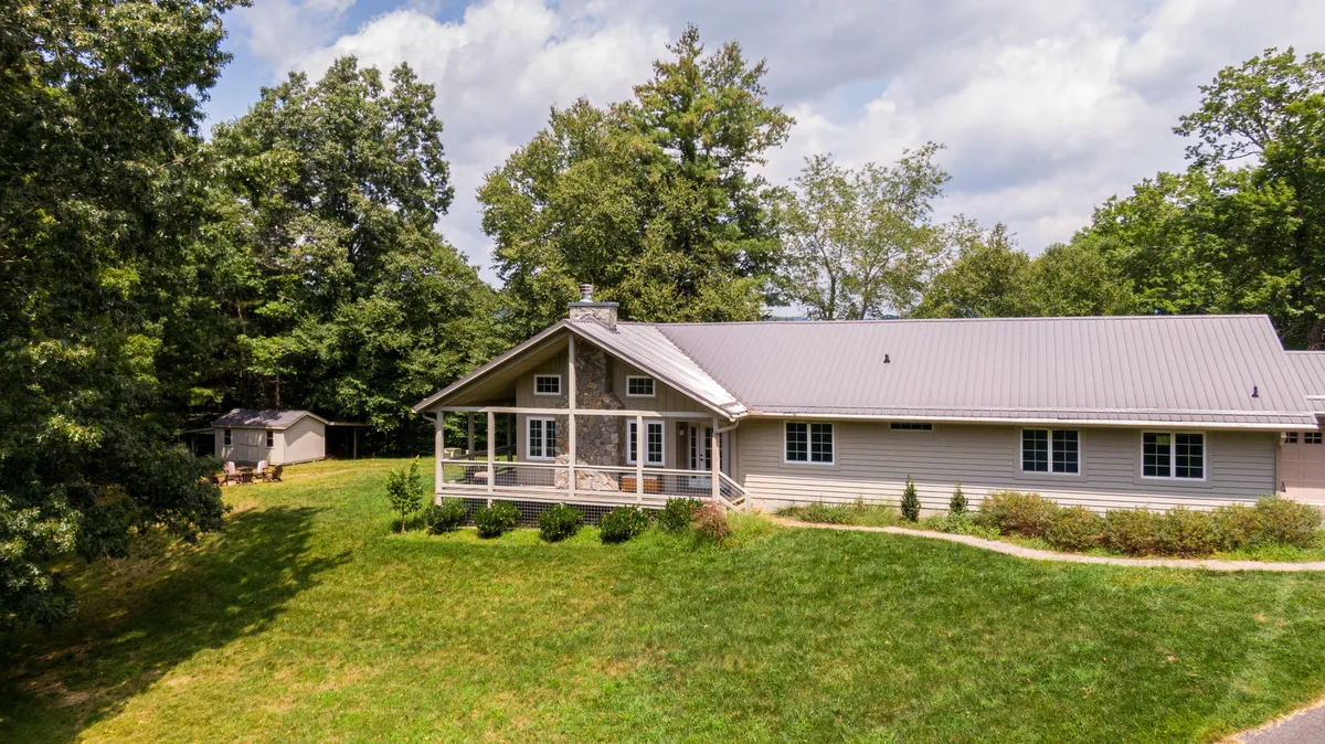 A view of the house with a porch, surrounded by lush greenery and a distant shed. The setting exudes a sense of welcome and possibility for exploration, with plenty of space for outdoor barbecues and nature-inspired activities for all ages.