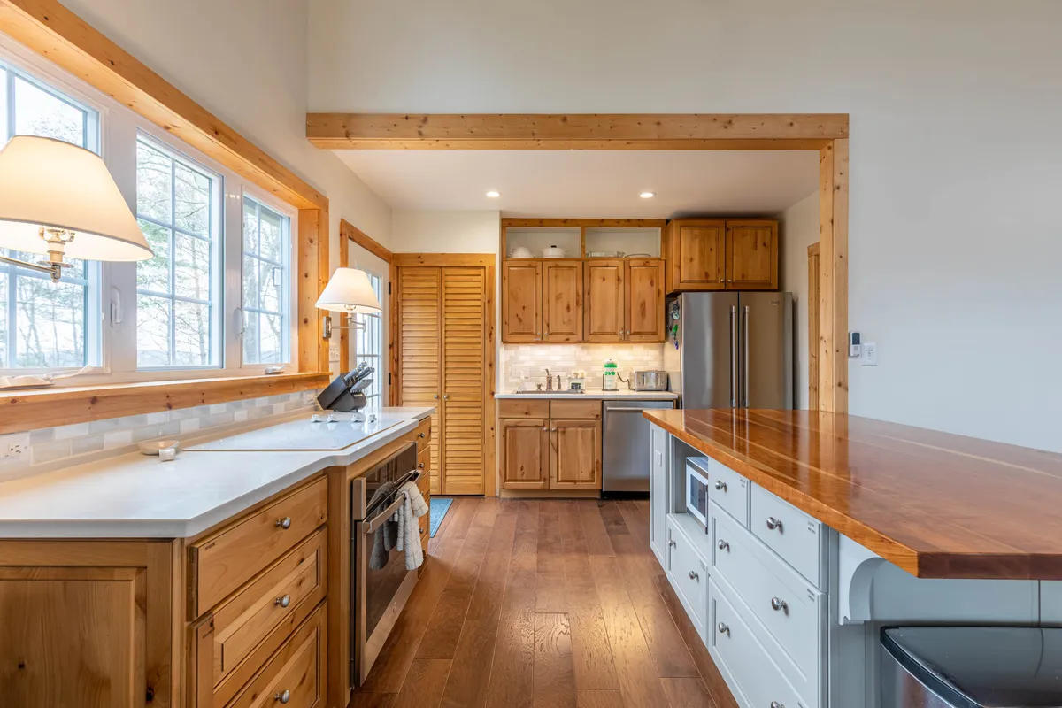 The focus is on a well-equipped kitchen with a prominent kitchen island, wooden cabinets, and expansive countertops. Ample natural light pours in through large windows, illuminating the cooking space. This setup hints at thoughtful amenities and space for culinary creativity.