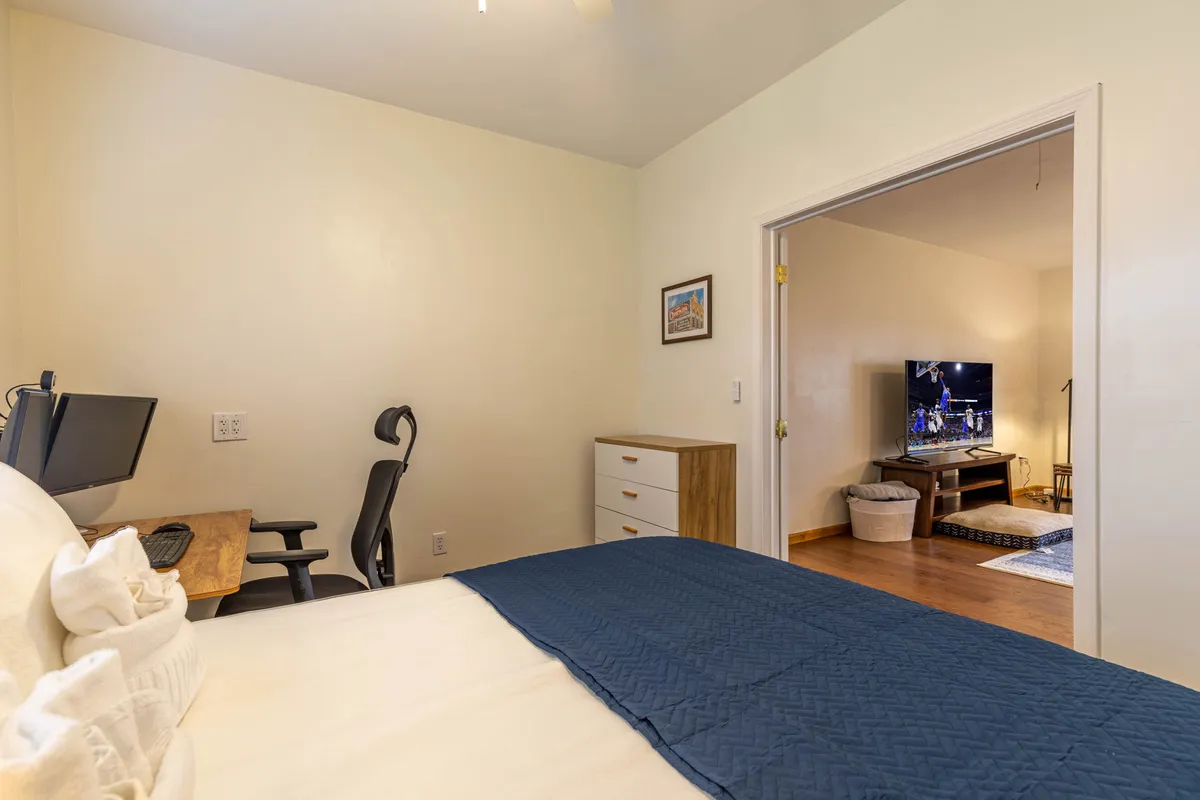 An inviting bedroom with blue bedding features a compact but highly functional computer setup, including a large monitor and desk, positioned for both work and entertainment. The adjacent room with a TV hints at the seamless transition between work and relaxation.