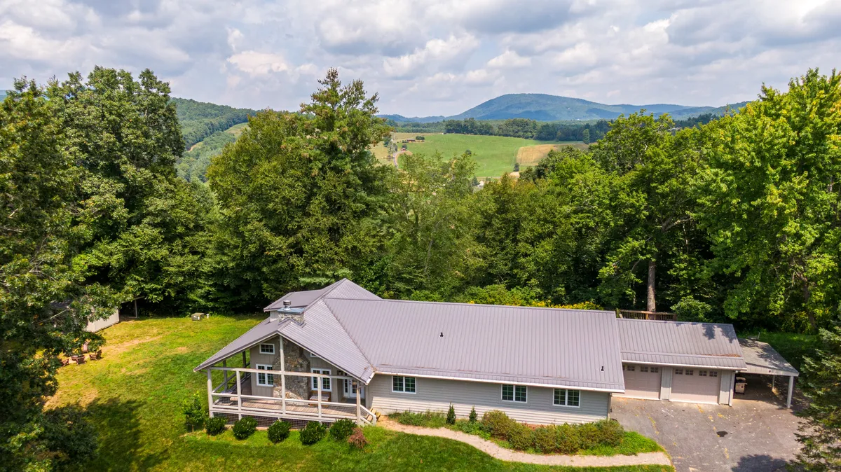 A wide exterior view of the River Refuge house nestled among lush greenery, with mountain peaks visible in the background. The inviting porch and expansive yard set a tranquil scene, suggesting plenty of space to step outside and enjoy the natural surroundings.