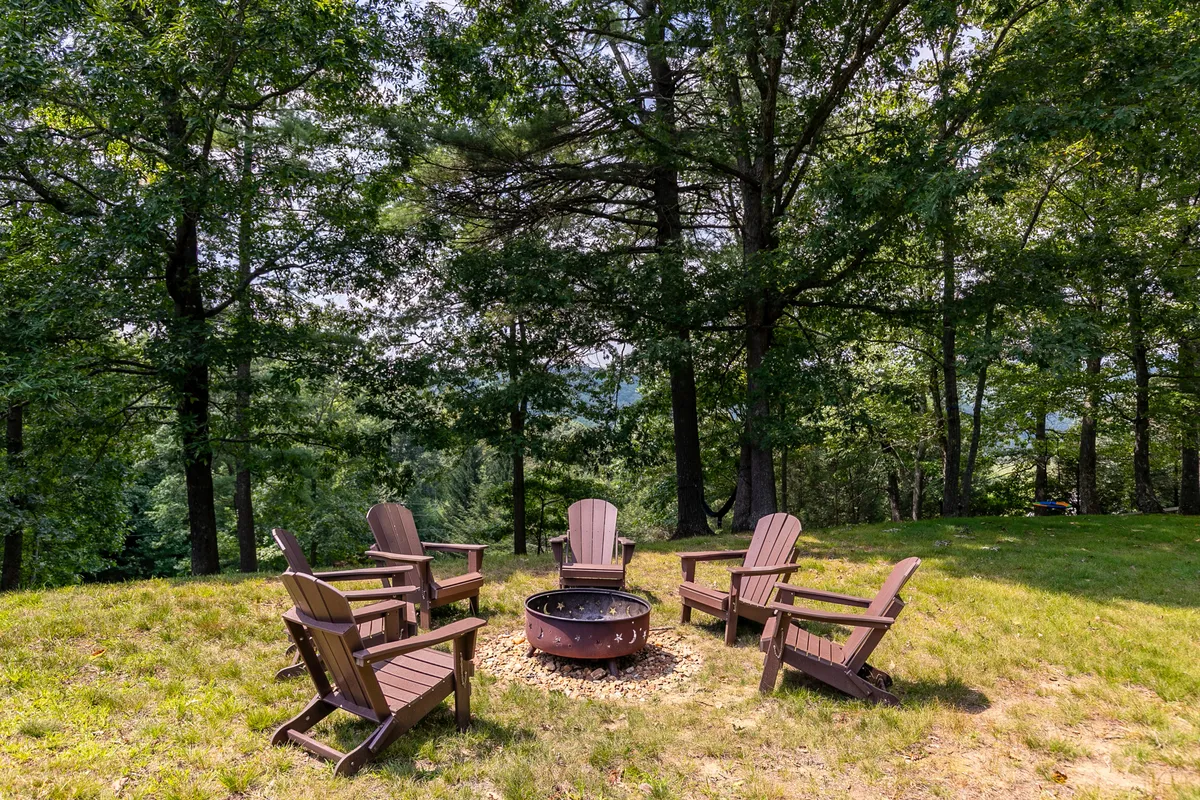 A welcoming fire pit surrounded by Adirondack chairs sits beneath tall trees, creating an inviting spot for families to gather. The setting suggests an environment where stories are shared, marshmallows are roasted, and children can experience the joy of nature just steps from the home.