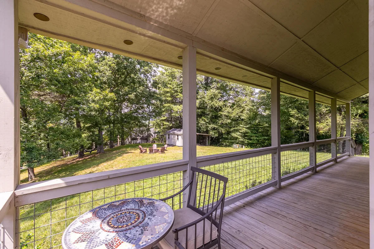 A peaceful outdoor seating area with a mosaic table and wooden chair, overlooking a green lawn and bordered by tall trees. The fire pit area is visible, enhancing the sense of a welcoming, social, and nature-connected environment.