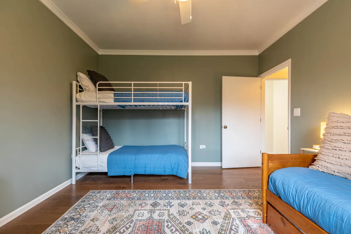 A bedroom designed for children, featuring a sturdy bunk bed alongside a single bed, topped with inviting bedding. The room includes a soft rug, nightstand, and a lamp, creating a playful yet cozy environment suitable for siblings or young guests. The arrangement encourages both rest and late-night chats, and the whimsical decor adds a touch of imagination.