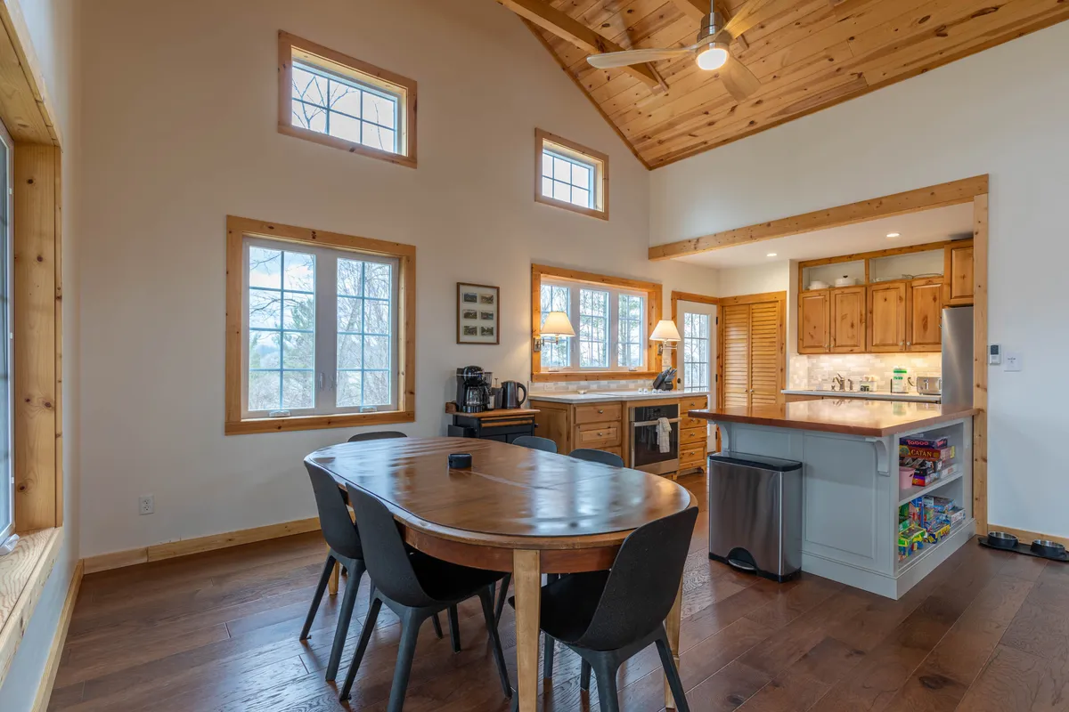 This image showcases a warm, welcoming dining area adjacent to a spacious kitchen island. The space is filled with natural light from generous windows, and features wooden cabinetry and a wooden ceiling that creates a cozy, communal atmosphere. The table is set up for family meals or gatherings, and the layout encourages conversation and togetherness.
