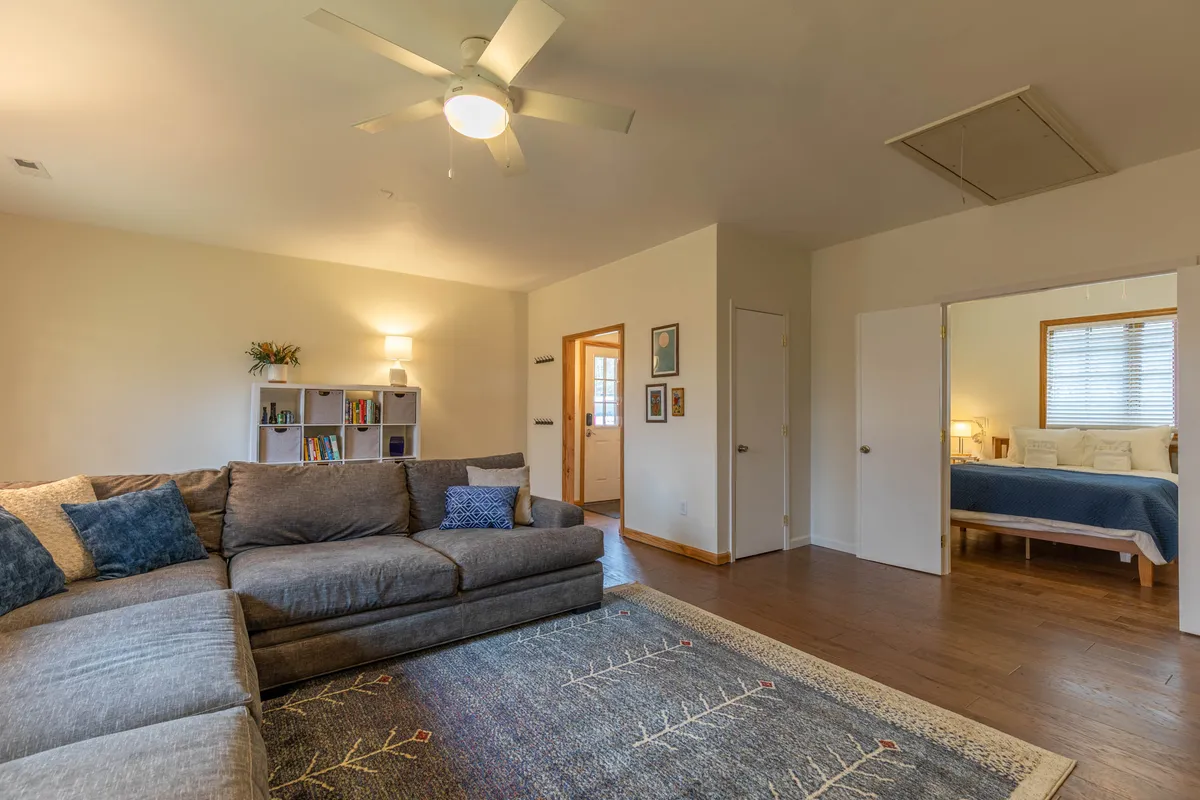 A cozy seating area with a comfortable sectional adorned with decorative cushions. Behind the sofa, a bookshelf filled with plants and books hints at areas for both leisure reading and games, while a stylish light fixture adds a warm ambiance.