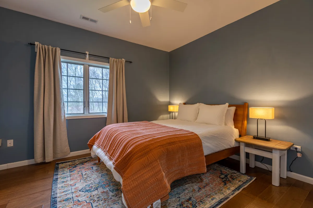 A serene bedroom with a comfortable bed dressed in soft linens and layered with an orange throw blanket, positioned near a window that allows natural light to gently illuminate the space. A small nightstand and lamp enhance the restful ambiance, while a patterned area rug adds warmth and texture.