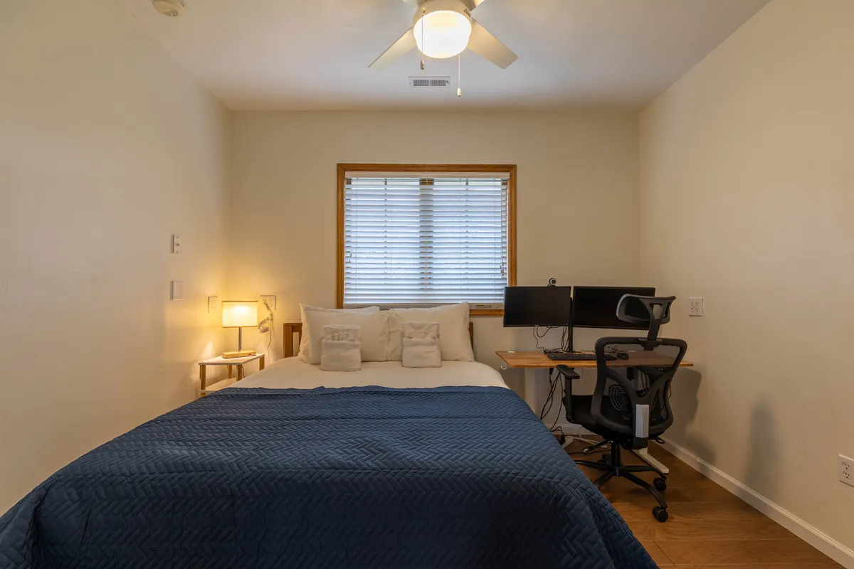 A cozy bedroom features a comfortable bed near a large window, with a dedicated desk setup that includes a monitor, task chair, and workspace essentials. The space is bathed in natural light, fostering both productivity and relaxation. Subtle decorative elements create a welcoming, homey atmosphere for work or study.