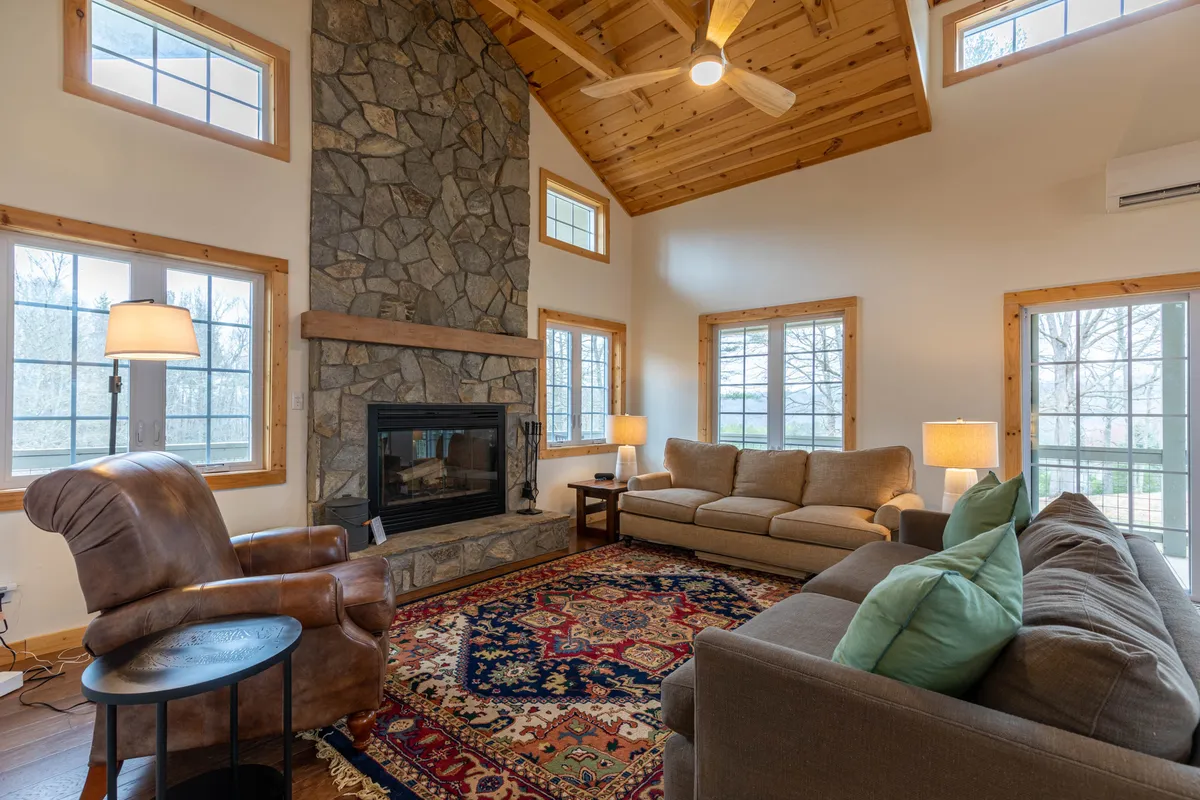 A sunlit great room with a vaulted ceiling, featuring a prominent stone fireplace as the central focal point. Comfortable seating is arranged around the hearth atop an ornate rug, while large windows flood the space with natural light and offer expansive views of the countryside.