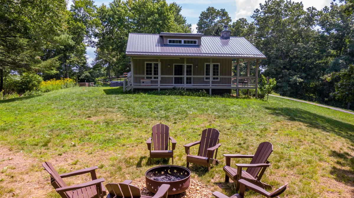 This exterior image features the house with a cozy fire pit surrounded by Adirondack chairs and lush greenery. The setting is perfect for evening gatherings, storytelling, stargazing, and enjoying the peaceful mountain environment.