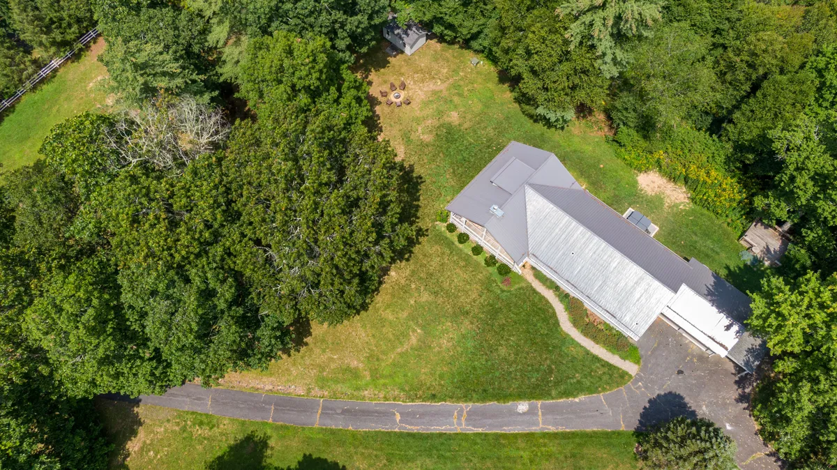 A wide view of the house set amid mature trees, with a visible driveway and a spacious yard featuring a cozy fire pit area. The property is surrounded by natural greenery, conveying a sense of peaceful seclusion and abundant outdoor space.